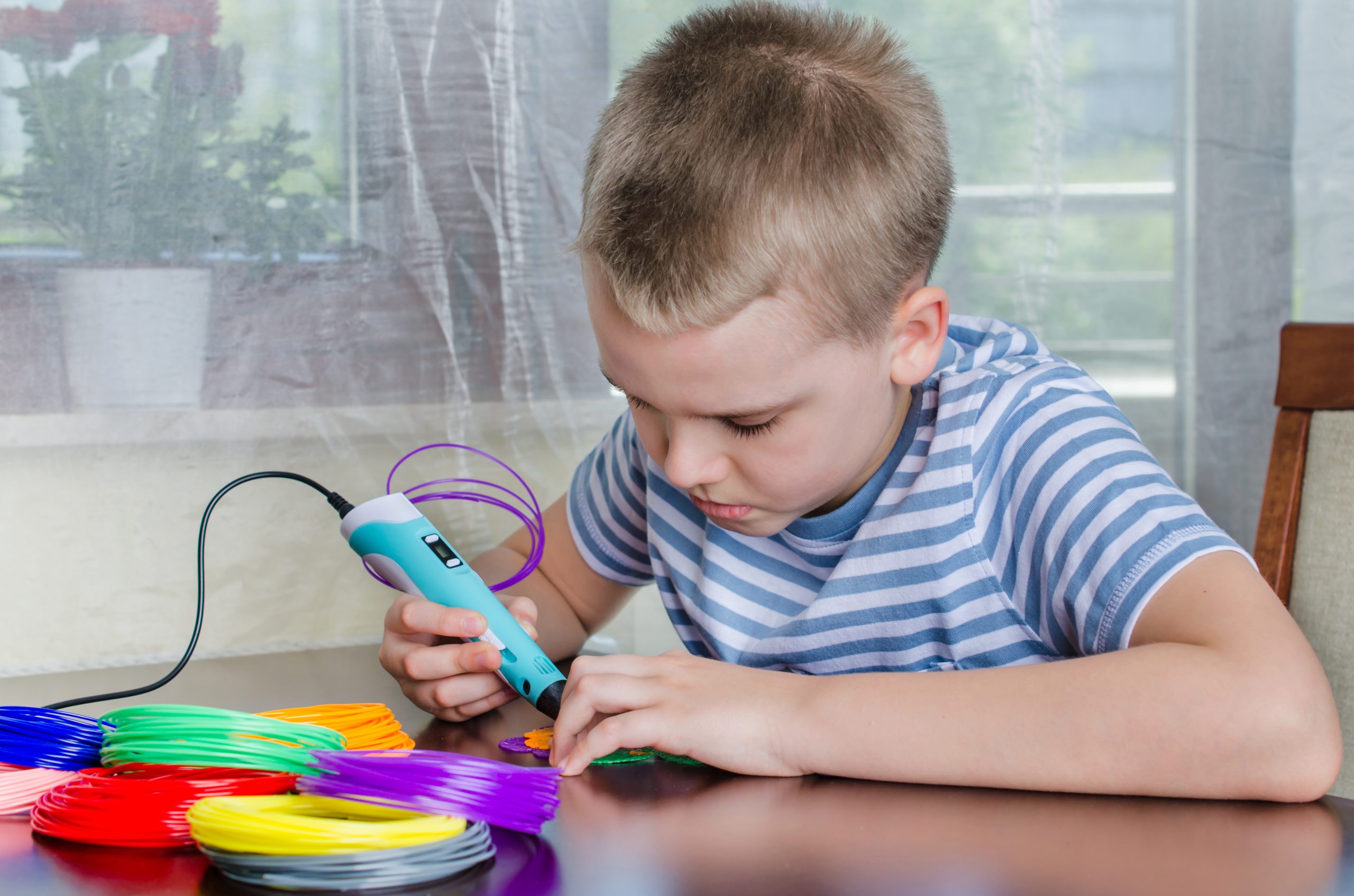 Boy using 3D pen. Happy child making flower from colored ABS plastic. Creative hobby at home, technology, leisure, education concept.